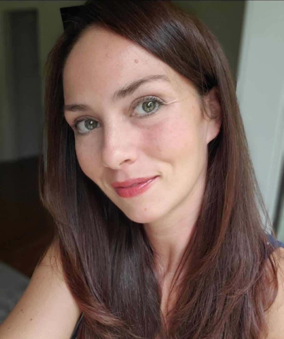 A woman with long brown hair, green eyes, and pale skin, smiling slightly toward the camera.